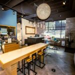 - Spa Bleü in West Dundee, St. Charles and South Barrington, IL Modern salon interior with product displays, a long wooden table with stools, a reception desk, and a large decorative chandelier, featuring brick walls and large windows—perfect for discovering Aveda Color and purchasing salon gift cards. - Spa Bleü in West Dundee, St. Charles and South Barrington, IL