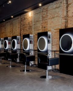 A row of modern salon stations with black chairs, circular mirrors with LED lights, and shelves against an exposed brick wall.
