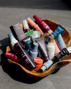 A wooden bowl filled with assorted bottles and tubes of Aveda hair and skincare products, arranged closely together on a gray surface.