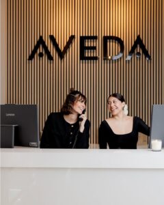 Two women are seated at a white reception desk in front of an "AVEDA" sign; one is on the phone and both are smiling.