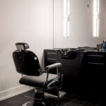 A black barber chair faces a mirror in a modern, empty barbershop with dark tiled floor and illuminated lights on the wall.
