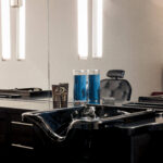 A black salon sink with blue disinfectant jars on the counter, modern lighting, and a salon chair reflected in the mirror.