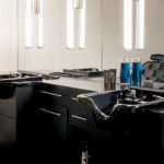 Modern salon wash station with two black shampoo sinks, a countertop, cabinets, large mirror, and two blue disinfectant jars.