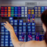 A person with tattoos organizes blue Aveda hair color boxes on a shelf, with other color-coded boxes and hair product tubes also visible.