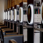 A row of black salon chairs faces mirrors with LED lights in a modern hair salon, with shelves of hair products between each station.