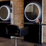 A modern salon station with a black styling chair, a round lighted mirror, hair products on a small table, and exposed brick walls in the background.