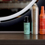 Four Aveda haircare products—Shampowder, Air Control, Nutri Plenish, and Color Control—are lined up on a wooden shelf beneath a curved mirror.