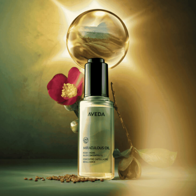A bottle of Aveda Miraculous Oil is displayed with a pink flower, seeds, and a glowing orb in the background.
