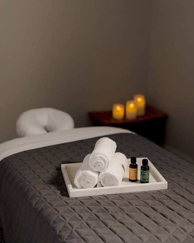 A massage table with a gray quilted cover holds a tray with rolled white towels and two small bottles. Lit candles are on a table in the background.