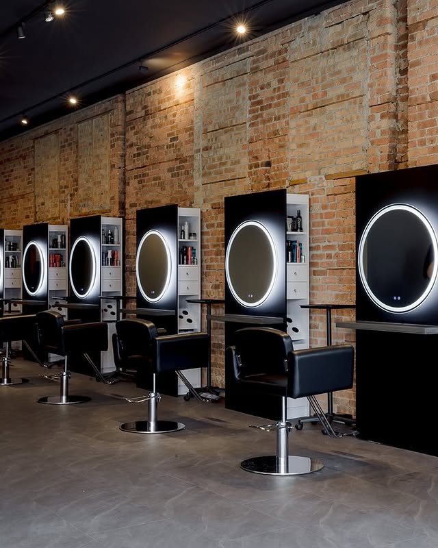A row of modern salon stations with black chairs, circular mirrors with LED lights, and shelves against an exposed brick wall.