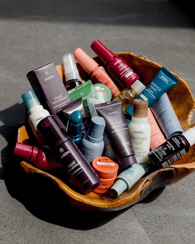 A wooden bowl filled with assorted bottles and tubes of Aveda hair and skincare products, arranged closely together on a gray surface.