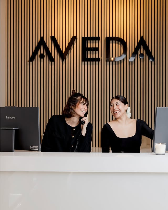 Two women are seated at a white reception desk in front of an "AVEDA" sign; one is on the phone and both are smiling.