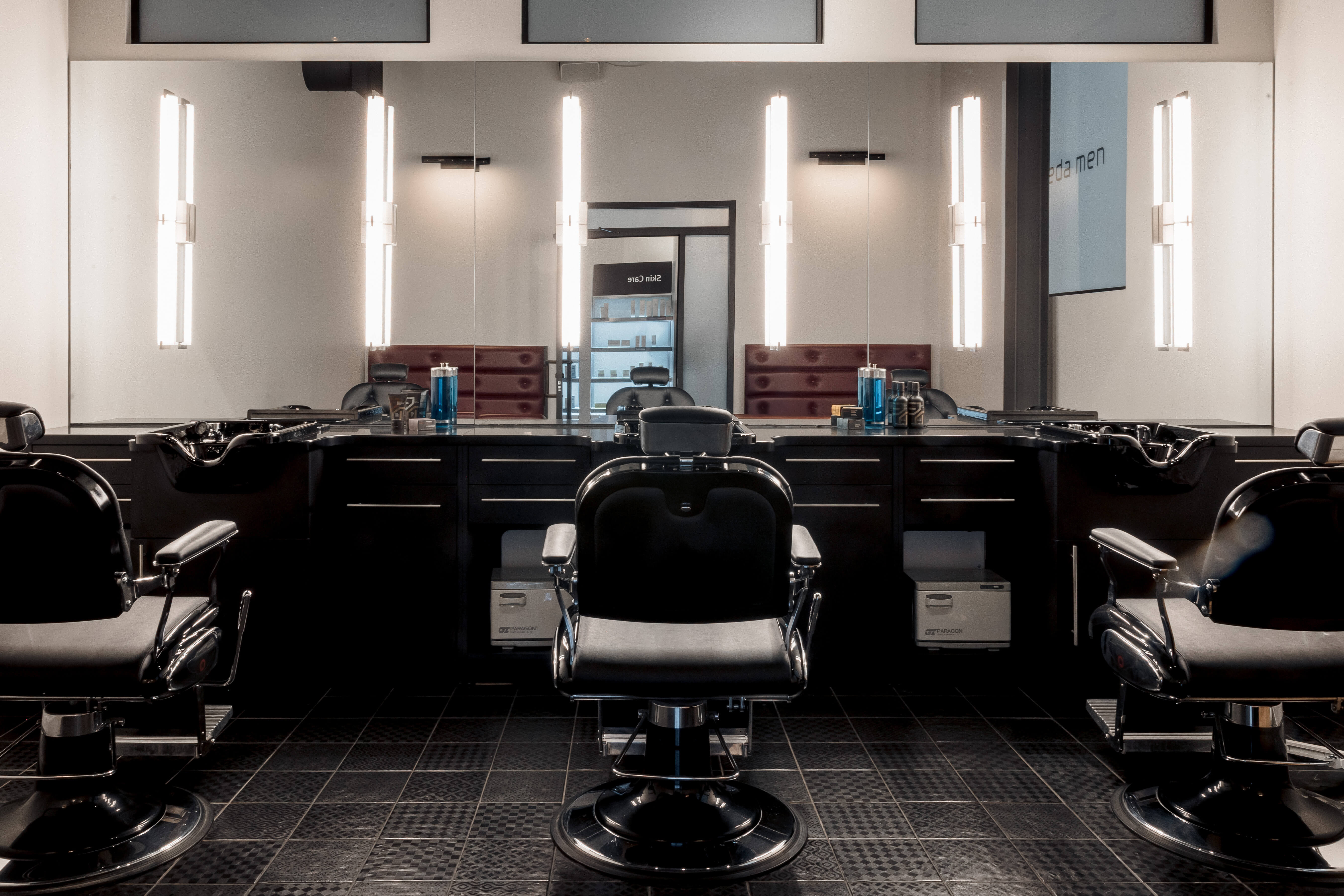 Three empty barber chairs face large mirrors in a modern, well-lit barbershop with dark cabinetry and grooming products on the counters.