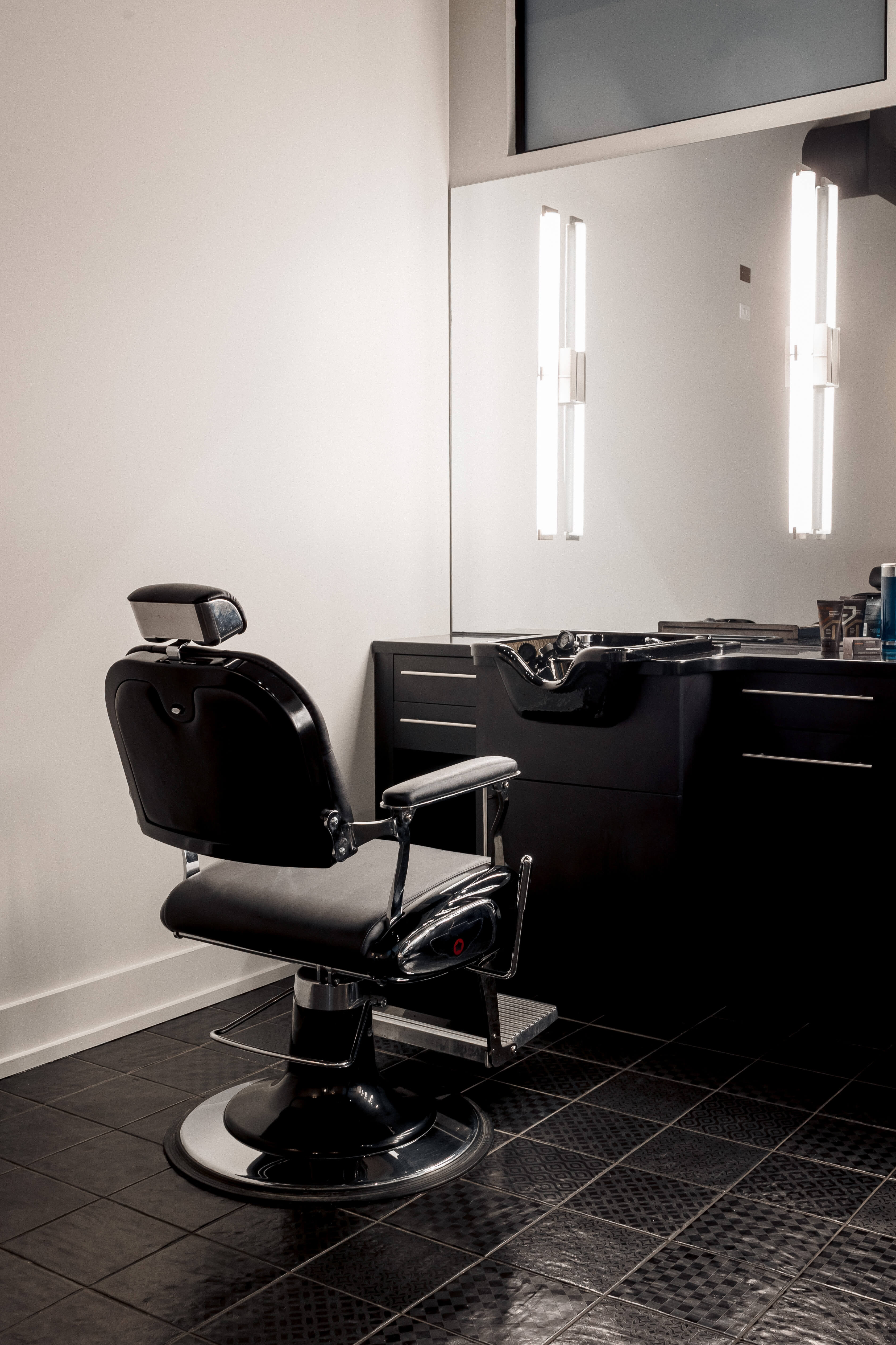 A black barber chair faces a mirror in a modern, empty barbershop with dark tiled floor and illuminated lights on the wall.