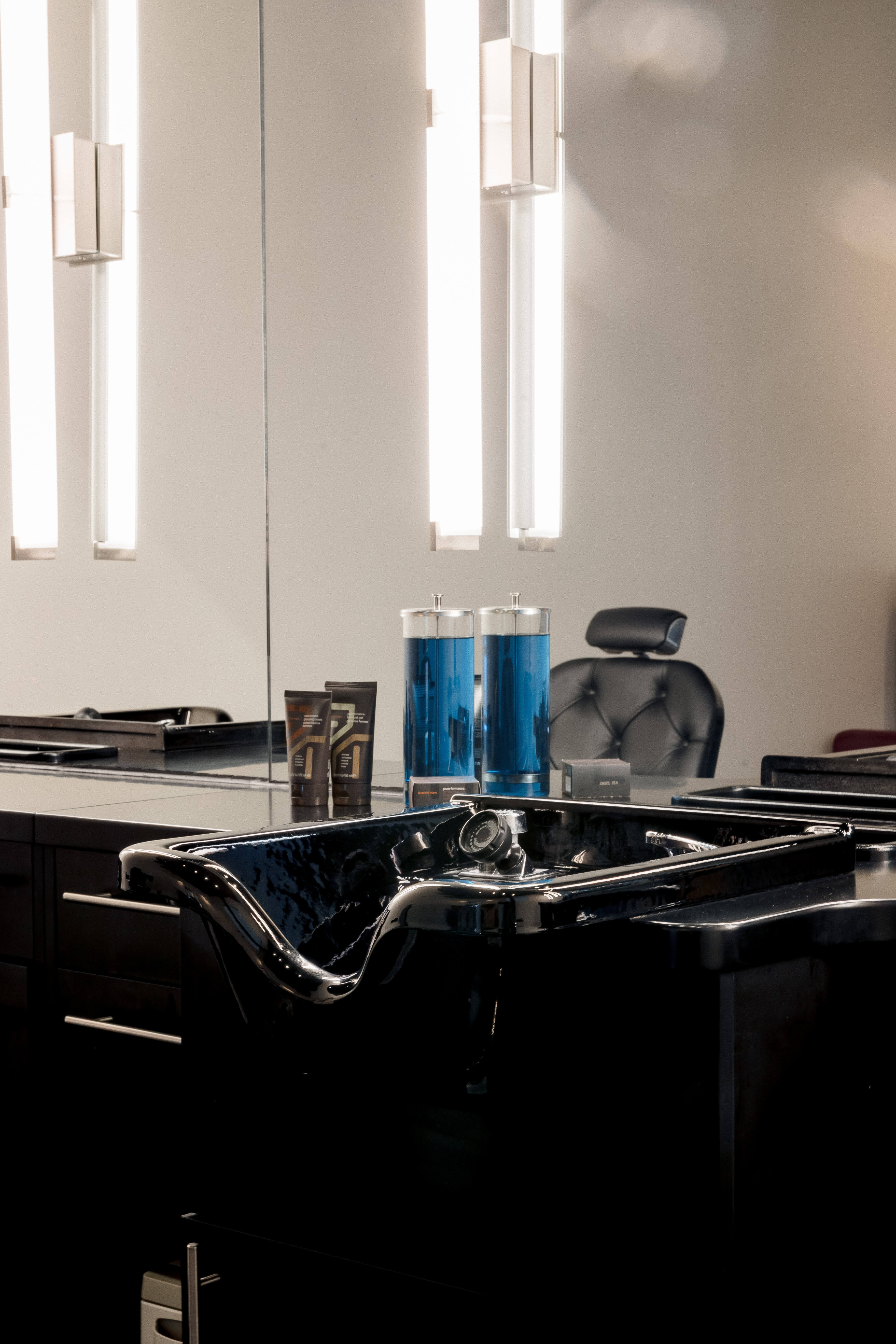 A black salon sink with blue disinfectant jars on the counter, modern lighting, and a salon chair reflected in the mirror.