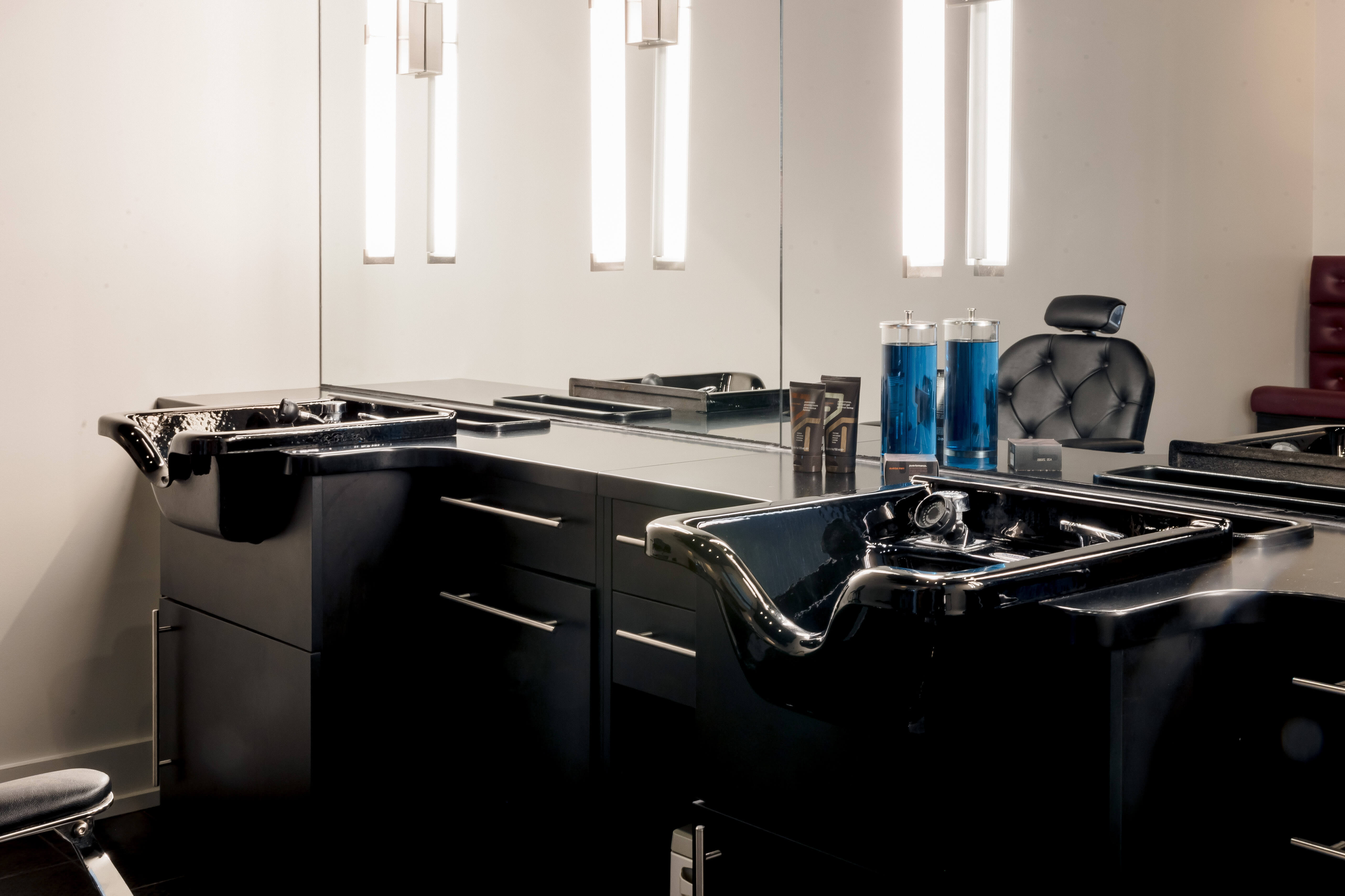 Modern salon wash station with two black shampoo sinks, a countertop, cabinets, large mirror, and two blue disinfectant jars.
