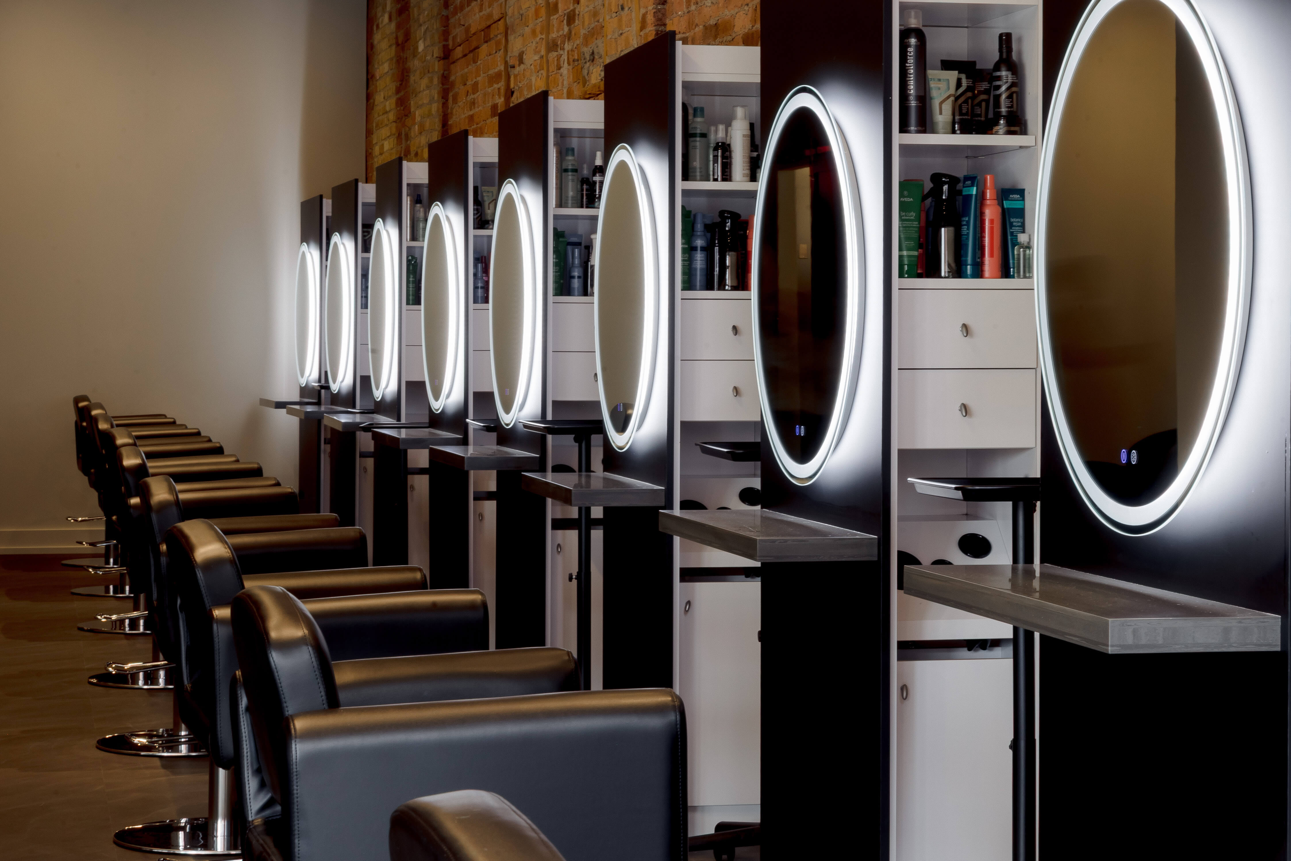 A row of black salon chairs faces mirrors with LED lights in a modern hair salon, with shelves of hair products between each station.