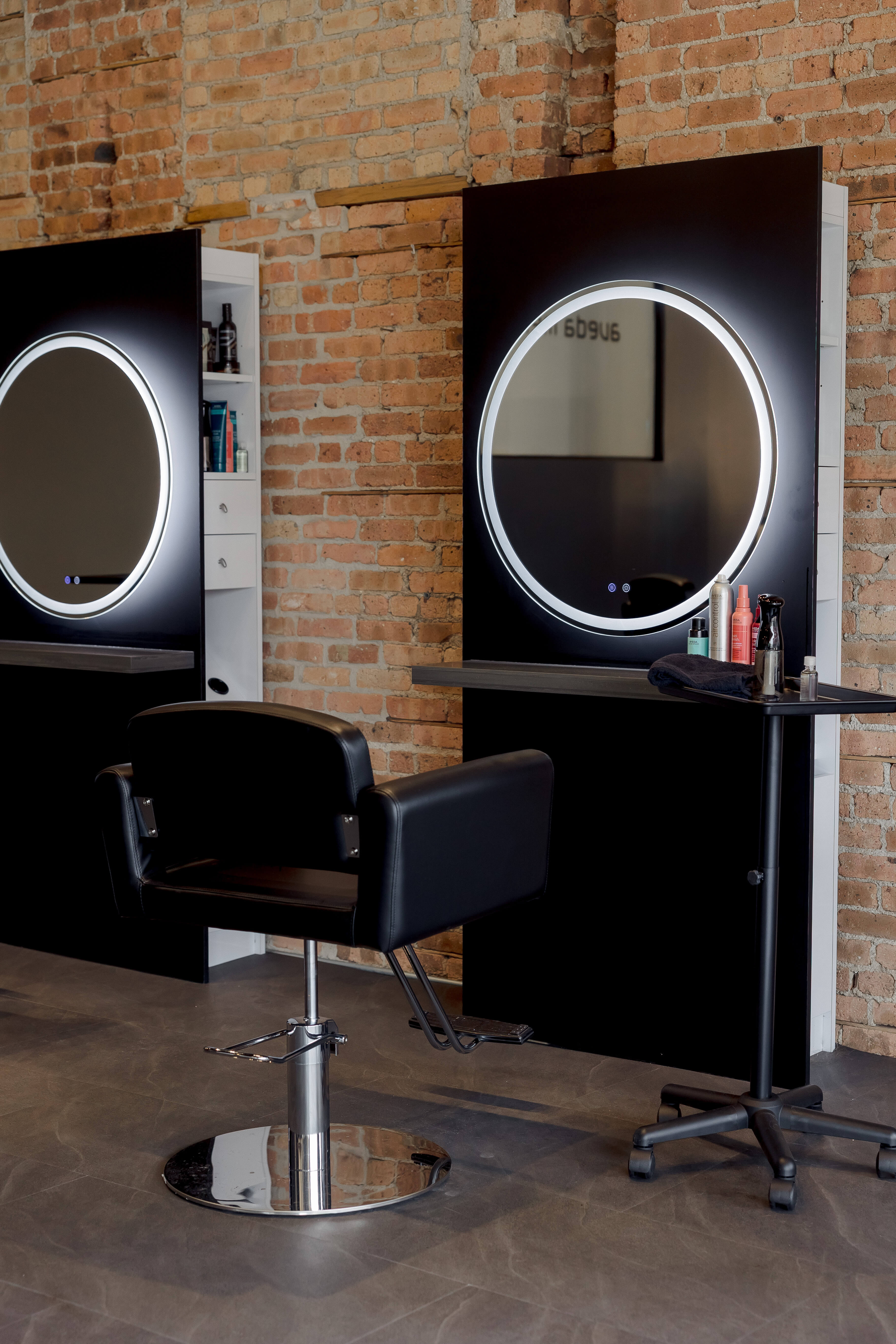 A modern salon station with a black styling chair, a round lighted mirror, hair products on a small table, and exposed brick walls in the background.