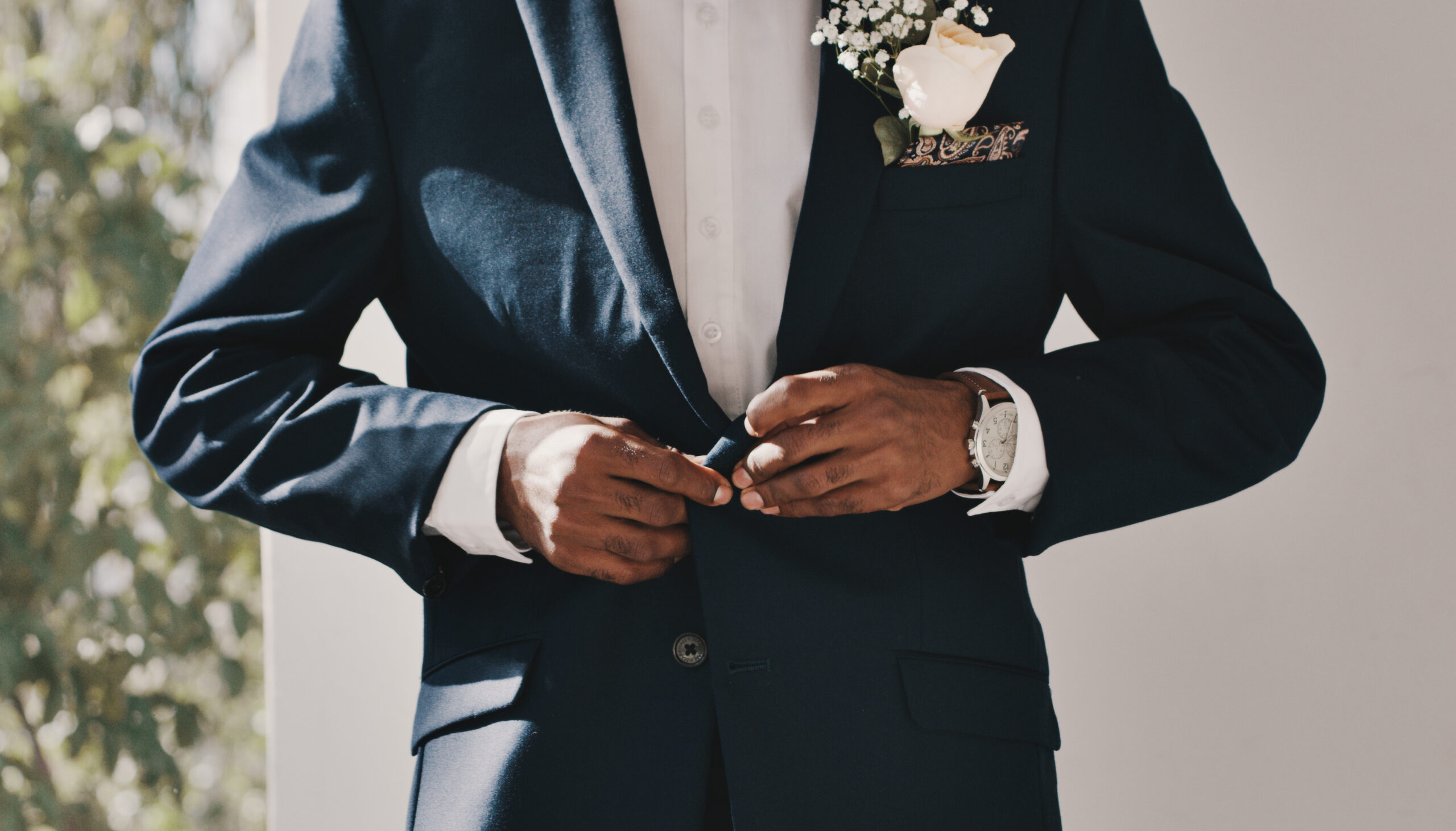 Man in a navy blue suit buttoning his jacket, wearing a white shirt, a watch, and a boutonniere with a white rose and baby's breath.